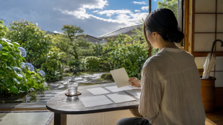 梅雨明け前後の日本の夏に、便箋を選びながら7月の時候の挨拶を考える女性