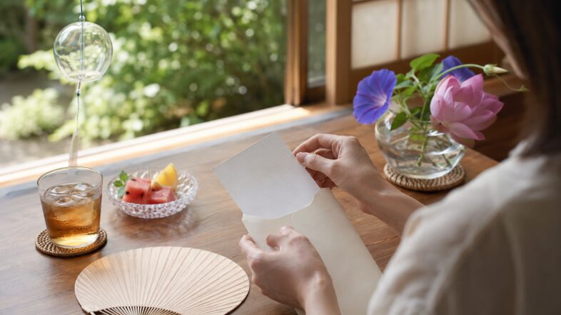 夏の窓辺で暑中見舞いのはがきを封筒に入れる、7月の挨拶状を表す画像