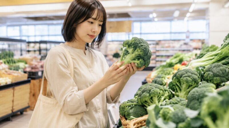 スーパーで日本人女性が新鮮なブロッコリーを選び、つぼみや茎を確認している画像